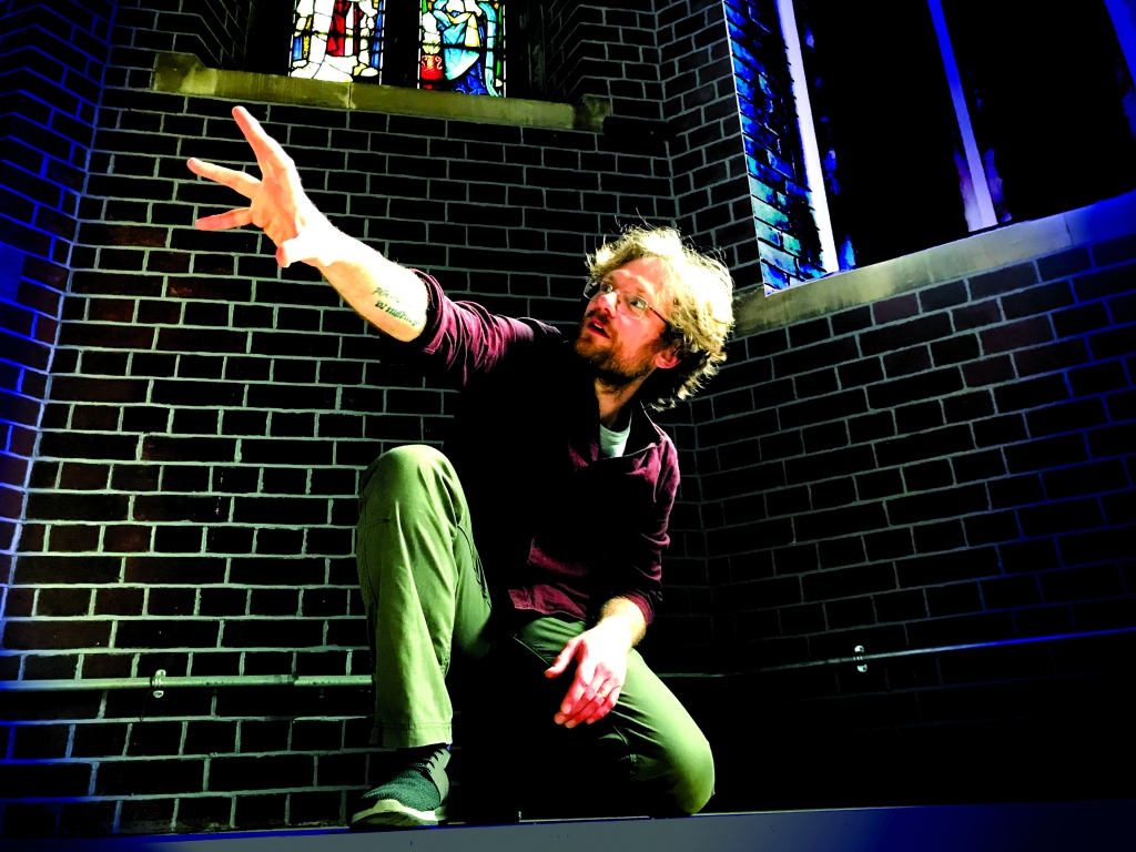 A man with wild hair and a short beard kneels on the floor in a church building, holding out one arm to the windows as he tells an audience a story.
